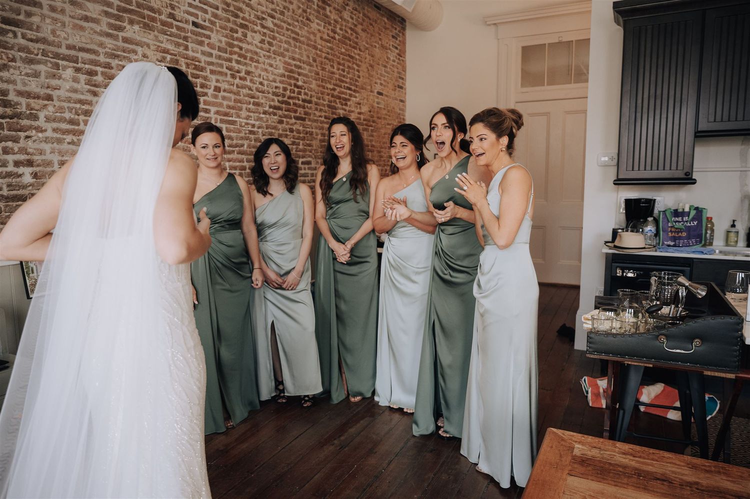 Bridal party in coordinated sage and silver dresses gather for formal moment in brick-walled room.