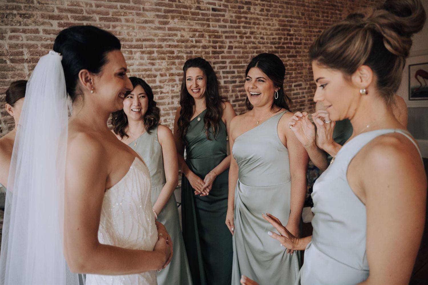 Wedding party members share candid reactions during bridal preparation in brick-walled room.