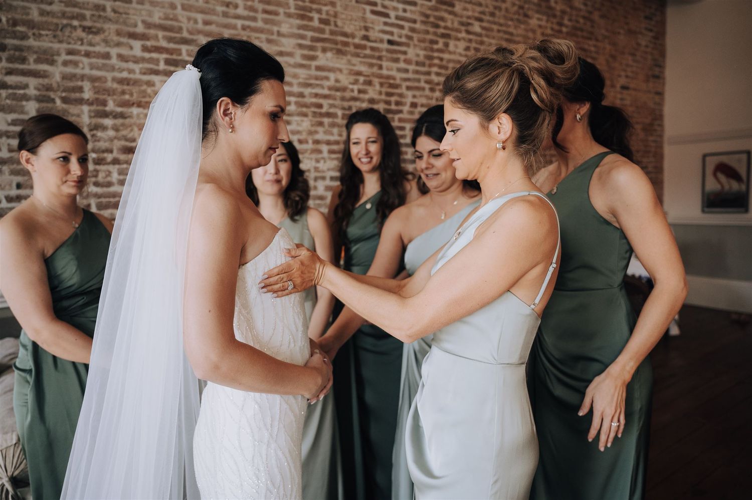 Emotional pre-wedding moment between bride in white gown and attendant in light-colored dress against brick backdrop.