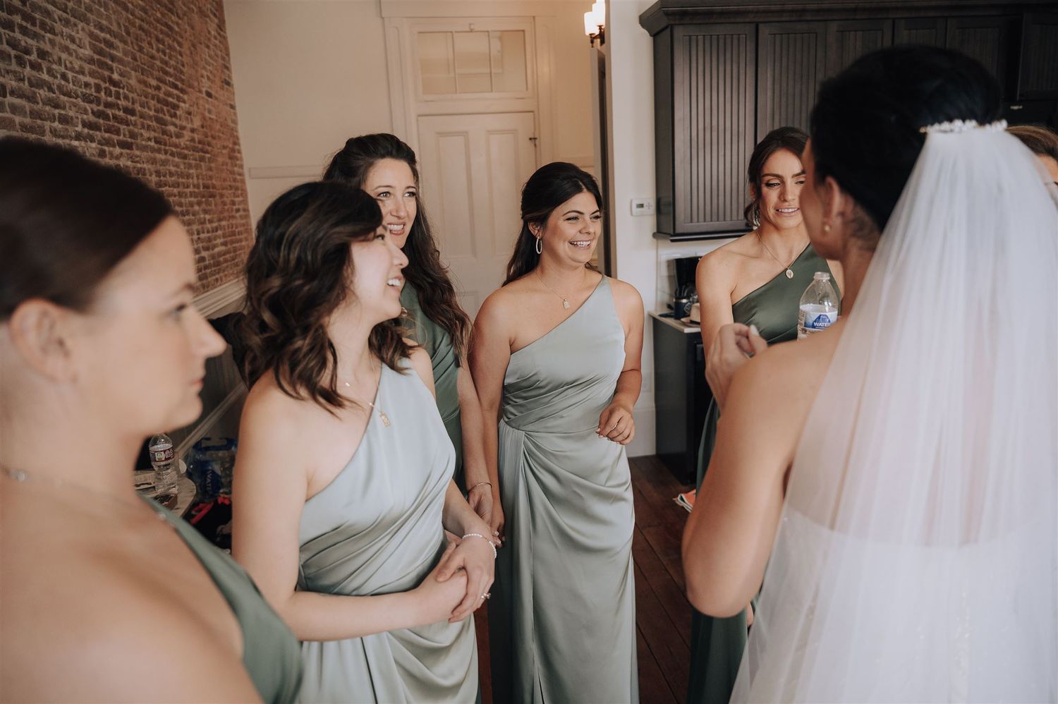 Bridesmaids in elegant sage gowns gather together during wedding preparations in front of exposed brick wall.