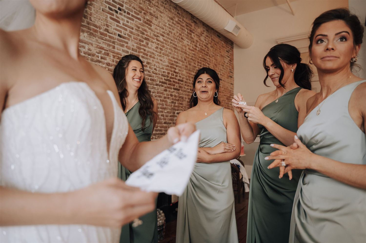 Bride in white dress shares a special moment with bridesmaids wearing sage green dresses in a rustic brick room.