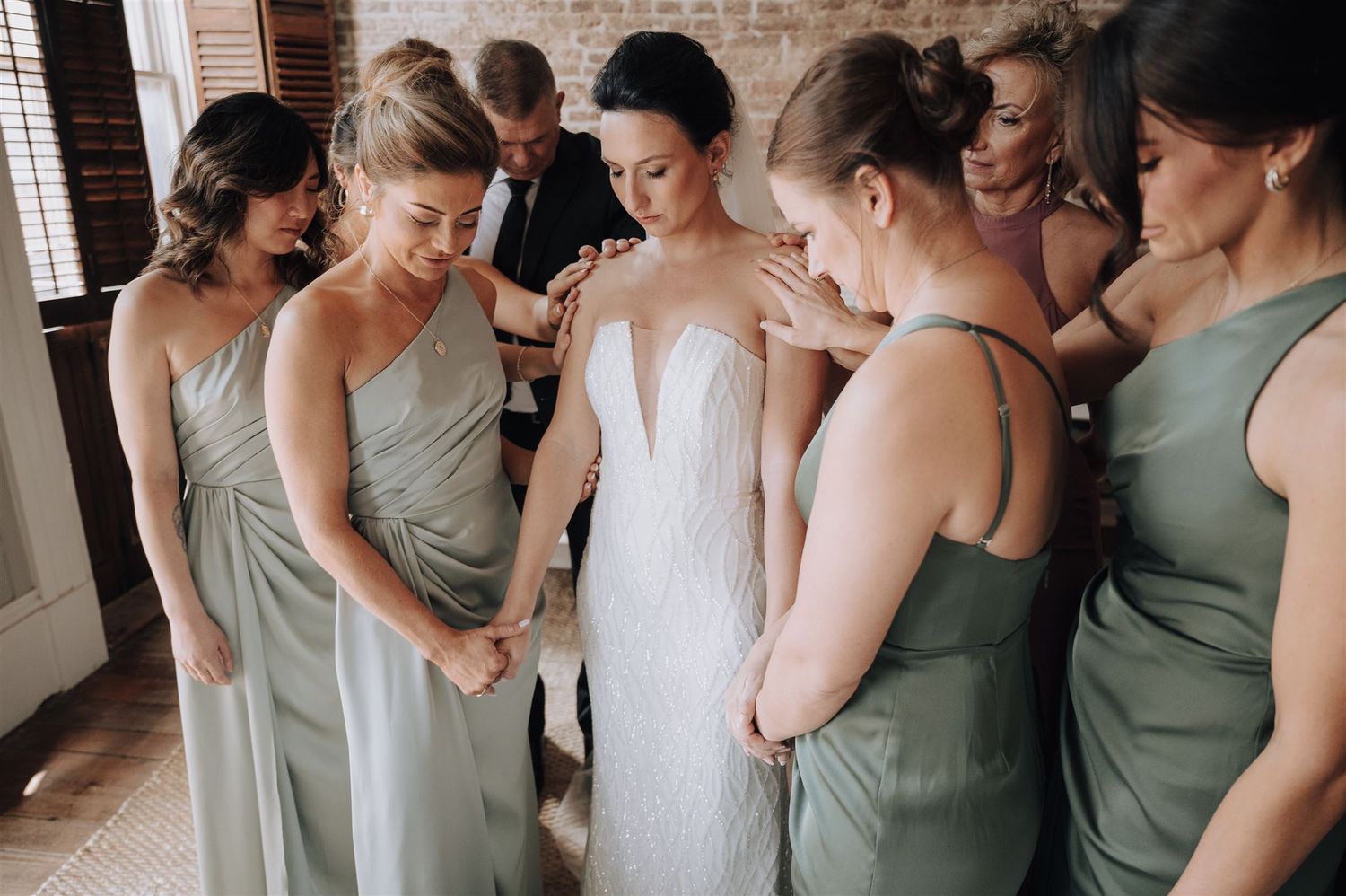 Wedding party gathers around in sage green dresses examining details of the white bridal gown.