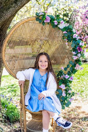 Outdoor photos of a child sitting on a wicker peacock chair surrounded by pink and white flowers.