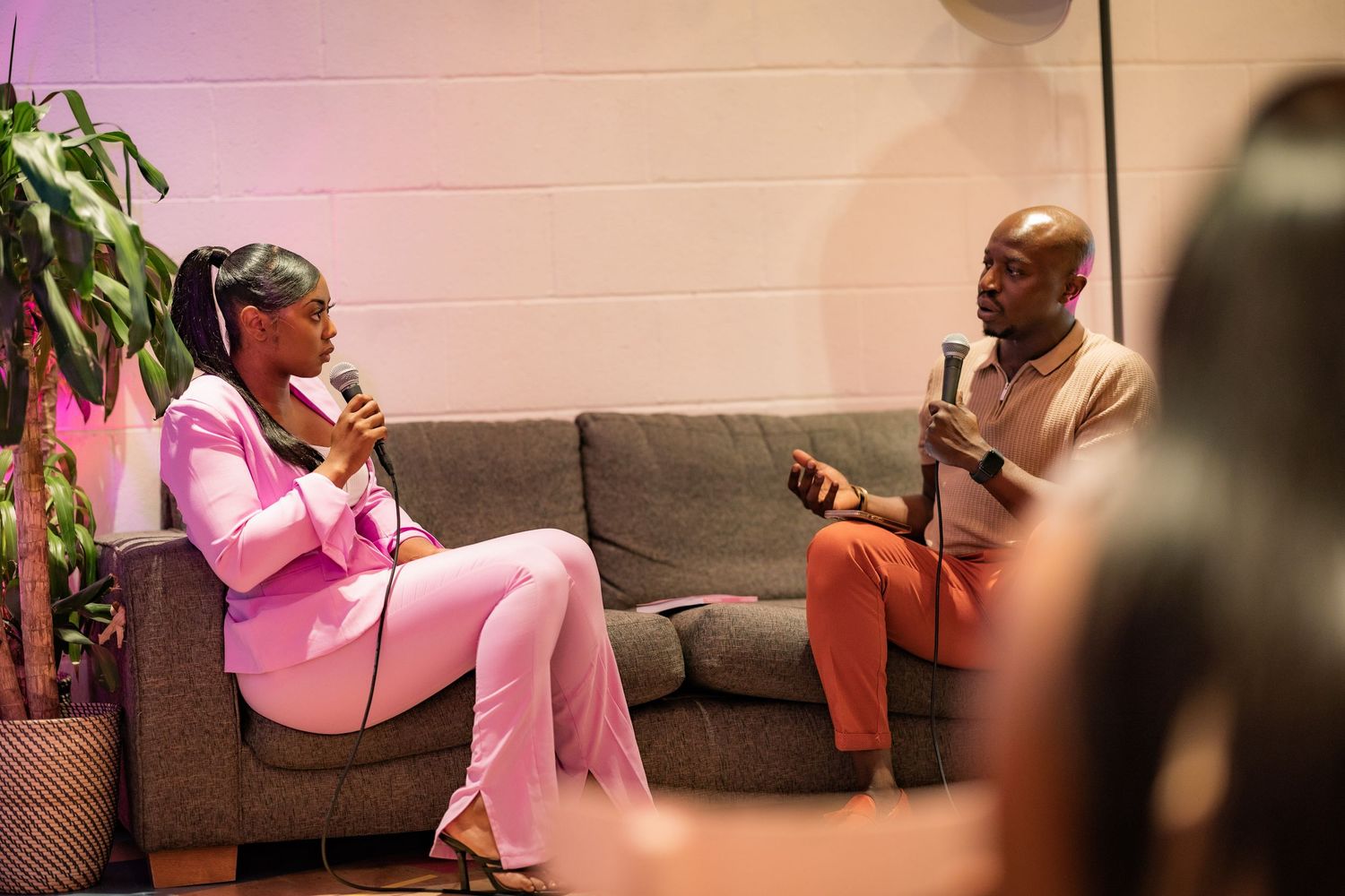 Two people engaged in a conversation while sitting on a gray couch with pink lighting.