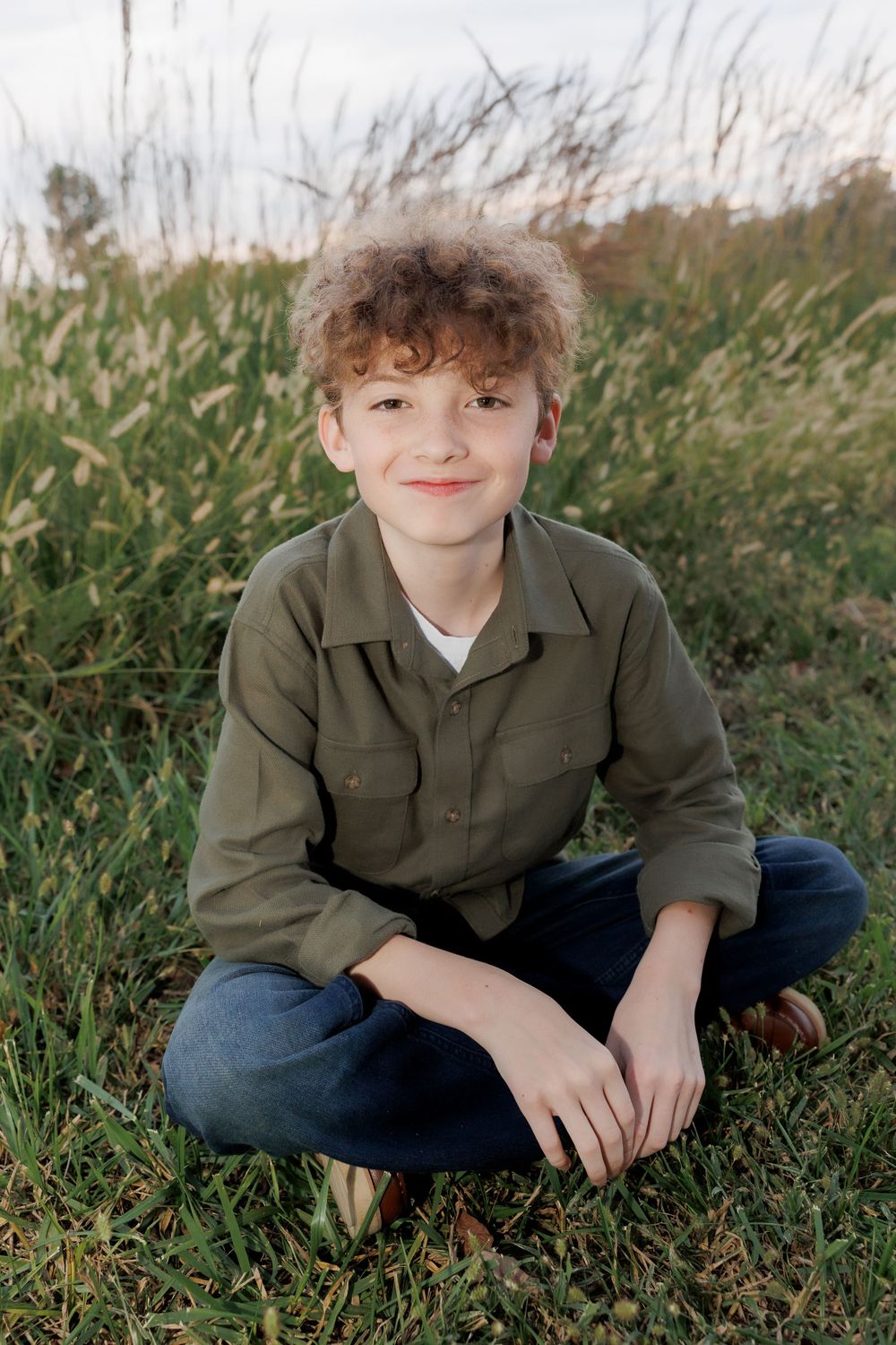 Youth sitting cross-legged in grass wearing olive jacket and jeans.