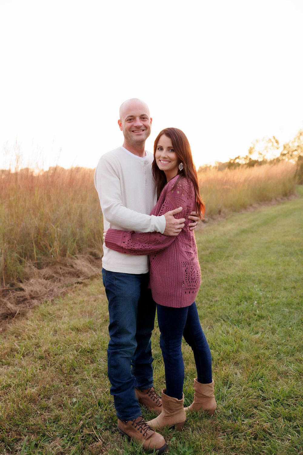 Couple in coordinating sweaters embrace while standing in a grassy field at sunset.
