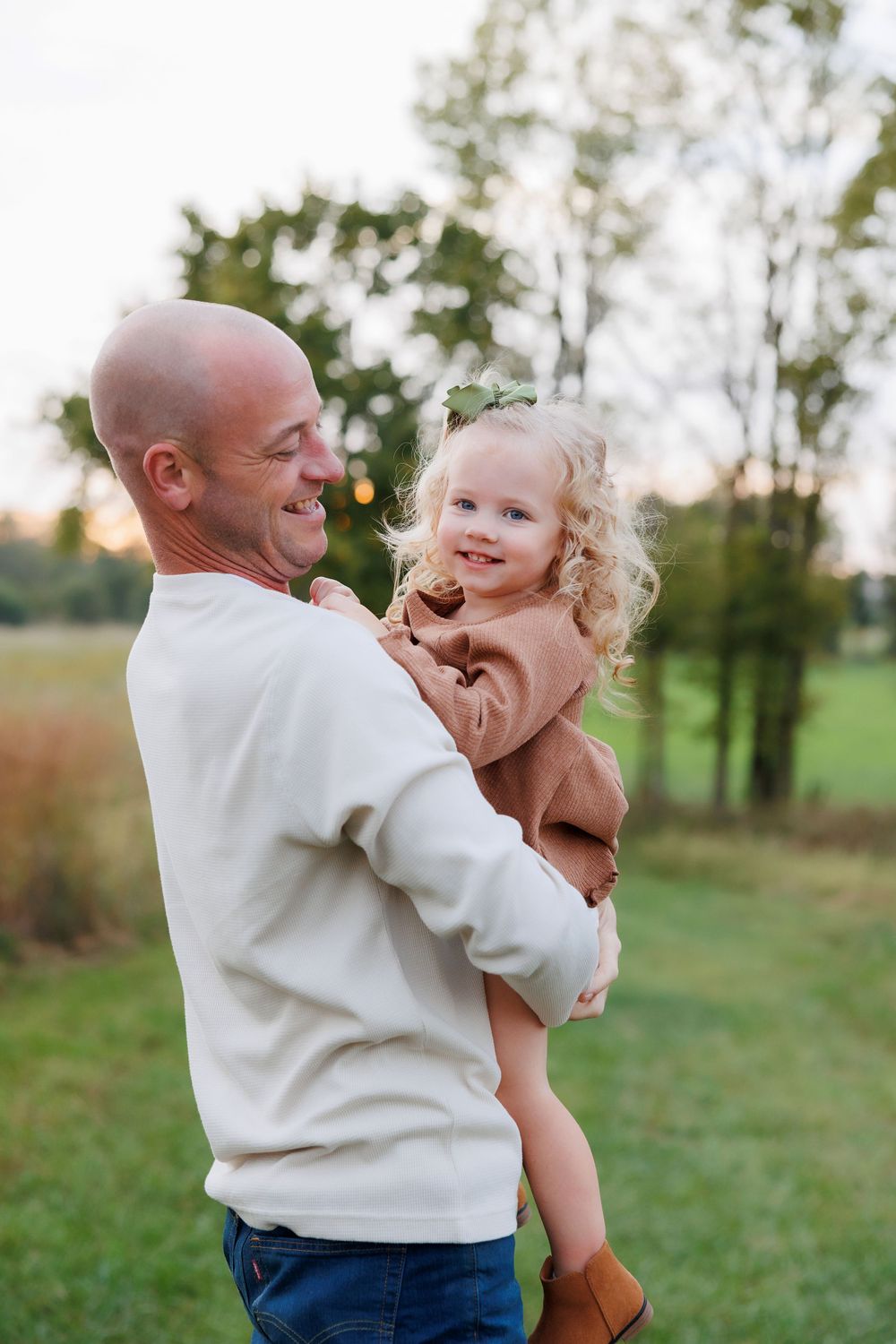 Adult in white sweater holds child in brown outfit while smiling outdoors.