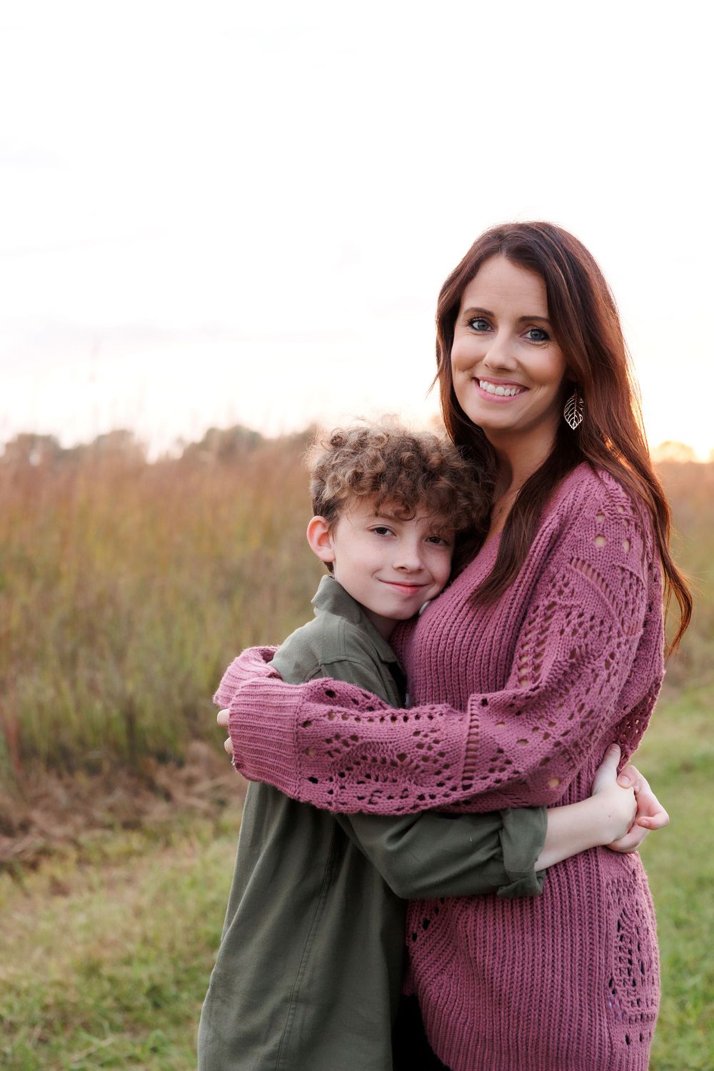 Two people embracing in field during golden hour wearing burgundy and olive clothing.
