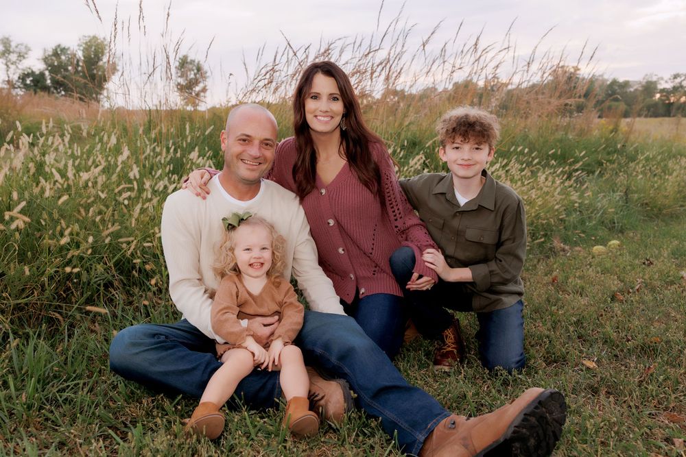 Family of four sits together on grass with tall wildflowers in the background.