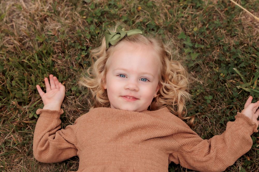 Young child with curly blonde hair lying on grass wearing brown sweater.