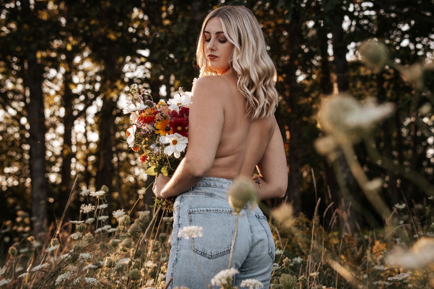 A woman in denim jeans holds autumn flowers while posing in golden sunlight.