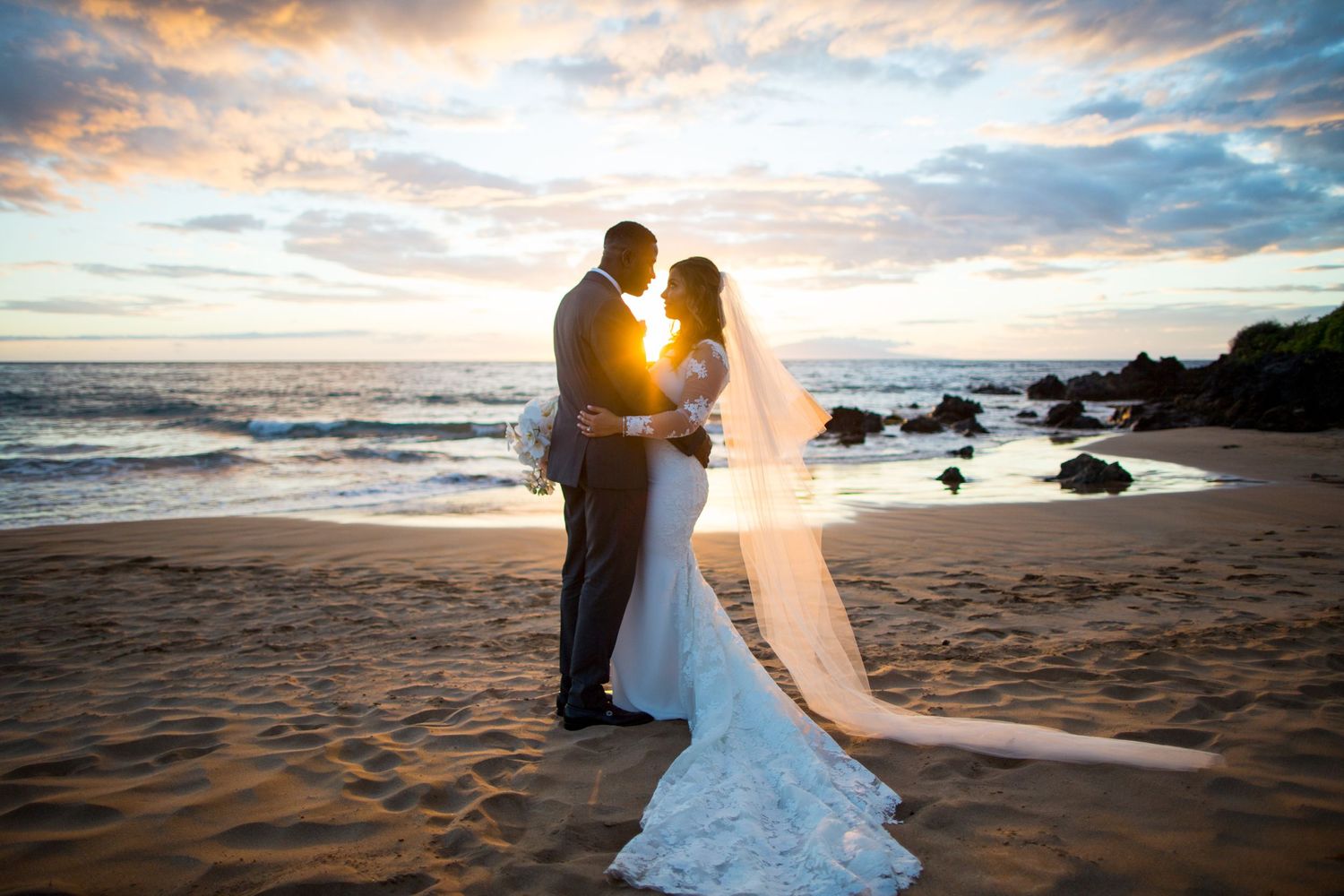 A couple shares a romantic moment on the beach at sunset with waves crashing behind them.