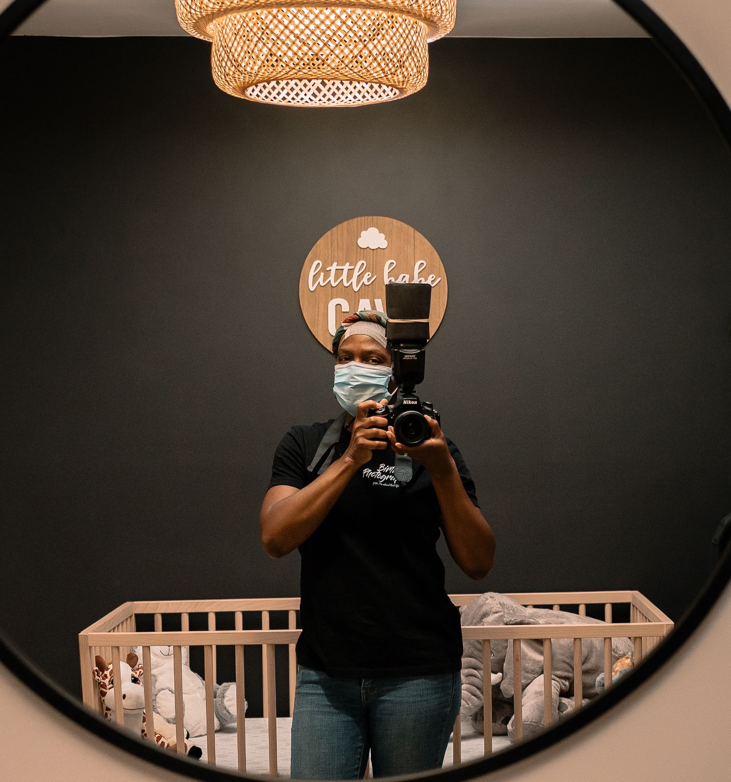 Black Birth Photographer Holding a camera wearing a mask standing in front of a baby crib - Joli Adore Photography