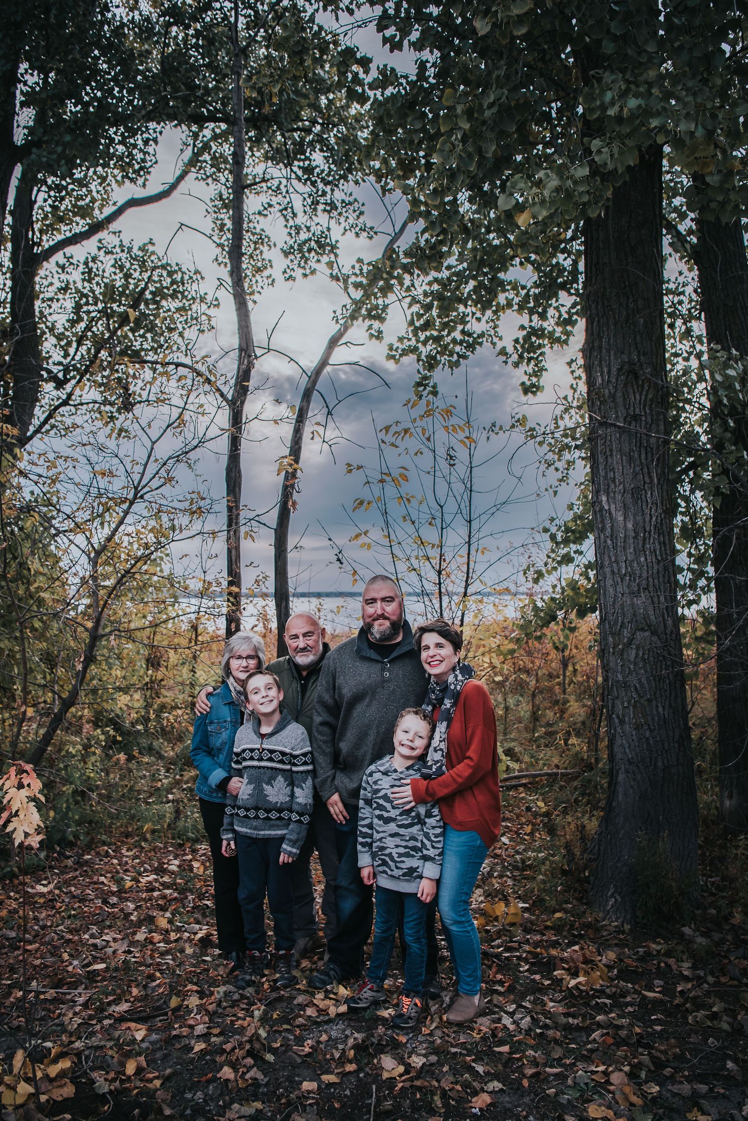 Extended family stand in a forested area with water and blue sky behind them at Brittania beach in the Fall