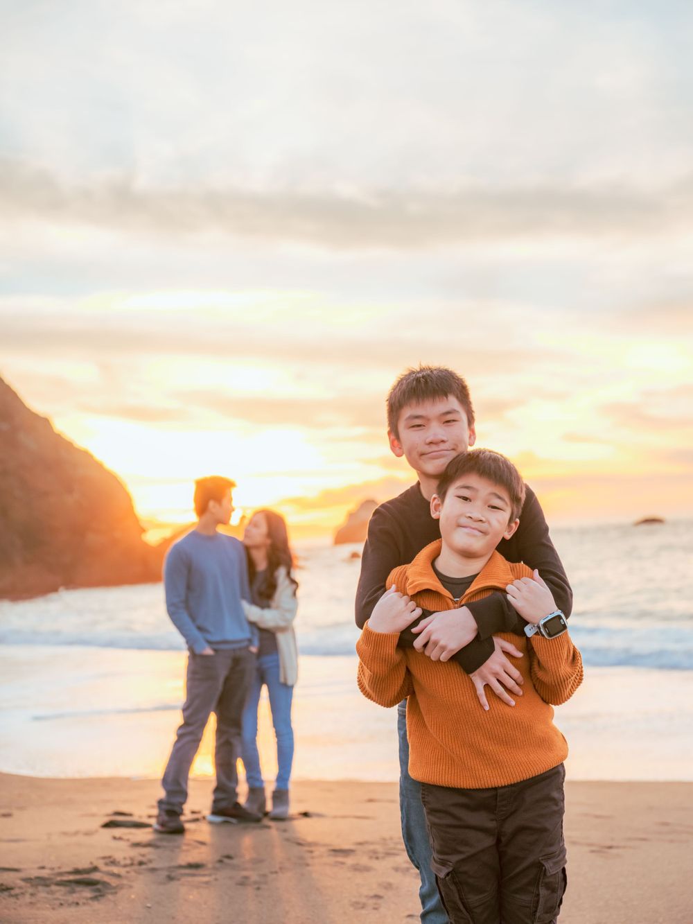 Two brothers hugging at sunset on a rocky beach while parents embrace in the golden background.