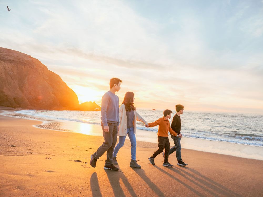 Family of four walking hand in hand along a beach at golden sunset near a rocky cliff.