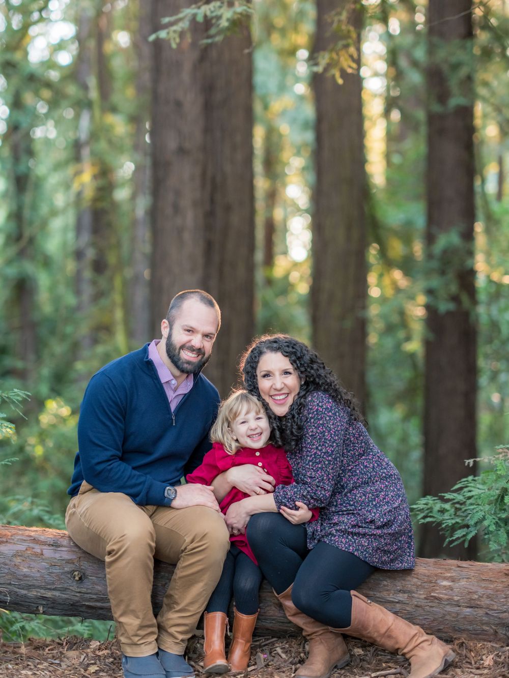 Smiling family of three posing on a log surrounded by tall redwood trees in a forest setting.