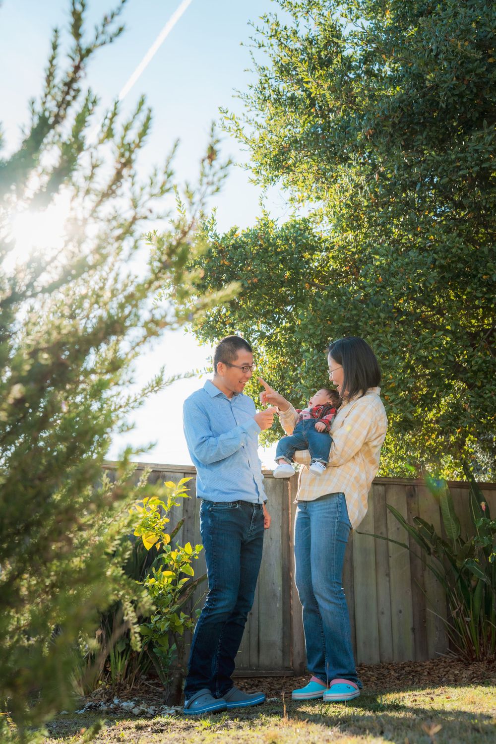 Asian couple holding their infant baby outdoors in a bright sunlit backyard garden.