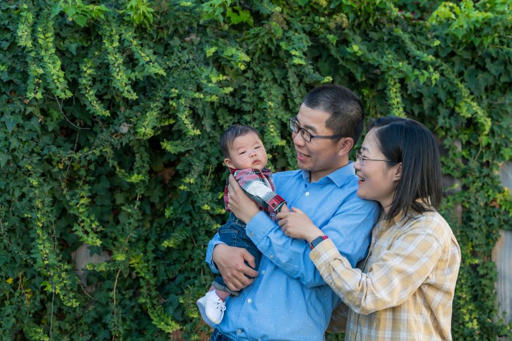 Asian family with parents holding their baby boy outdoors against a lush green ivy wall.