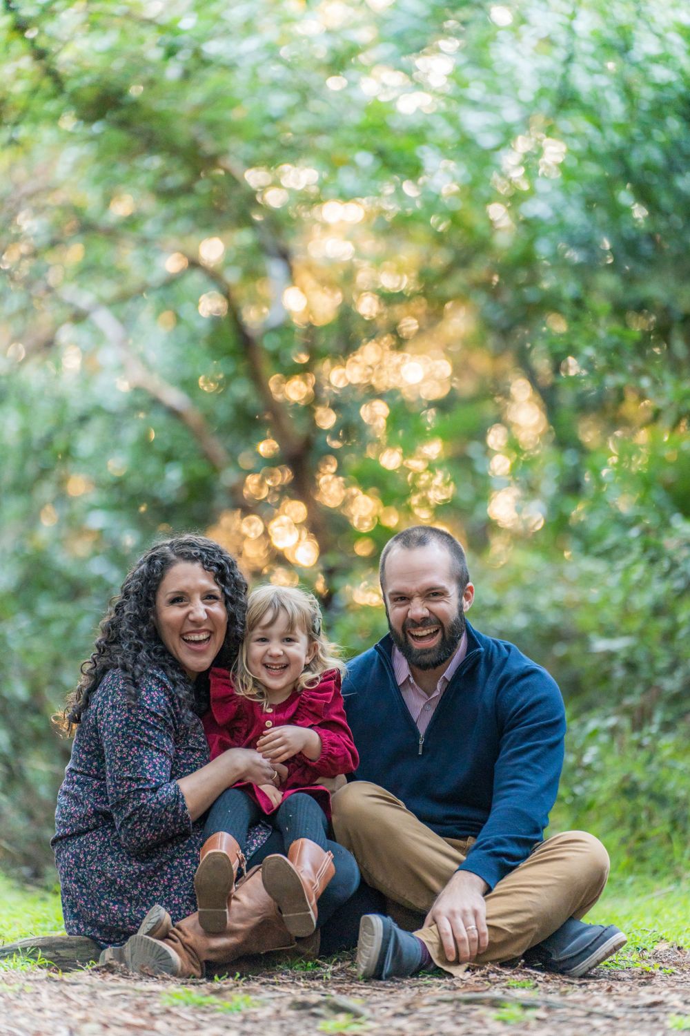 Laughing family of three sitting on the ground in a sun-dappled green forest, child in red dress.