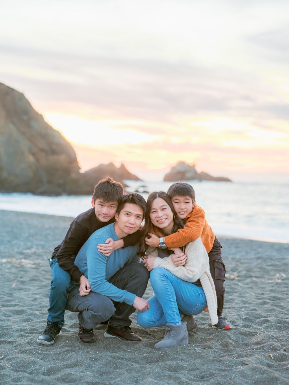Asian family of four smiling together on a sandy beach during a colorful sunset.
