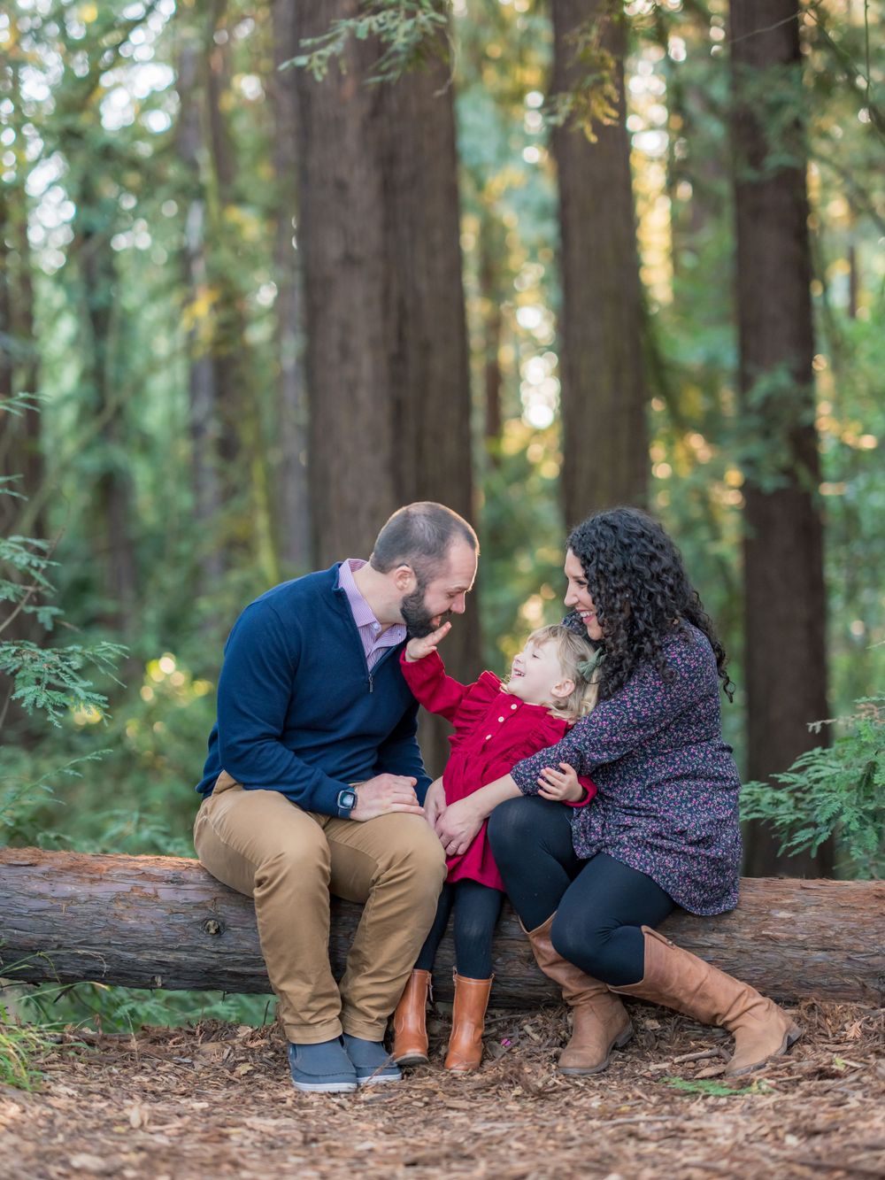 Family of three sitting on a fallen log in a redwood forest, parents smiling at toddler in red coat.