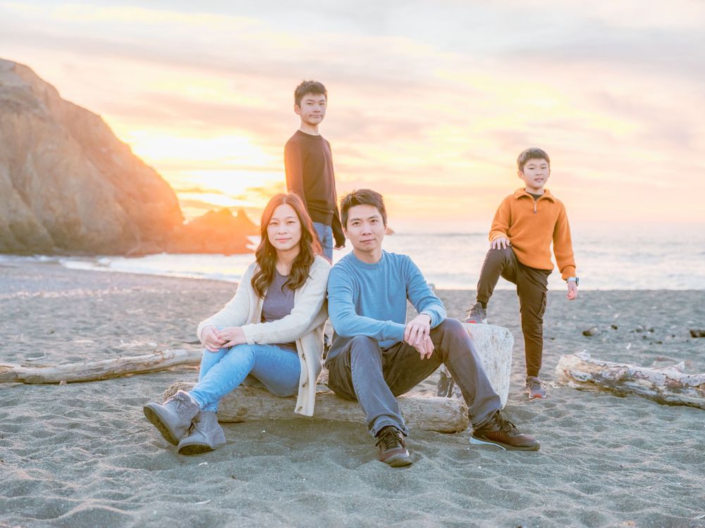 Family of four posing on driftwood at a dramatic beach sunset with rocky cliffs behind them.