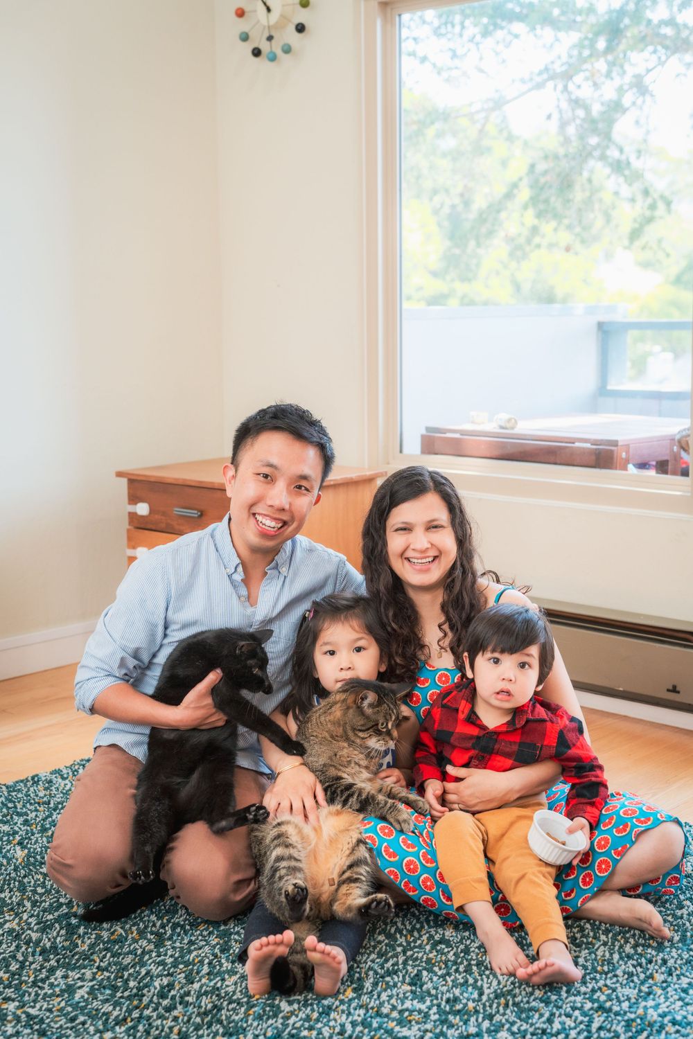 Family of four smiling indoors on a teal rug with a cat and puppy in a bright living room.