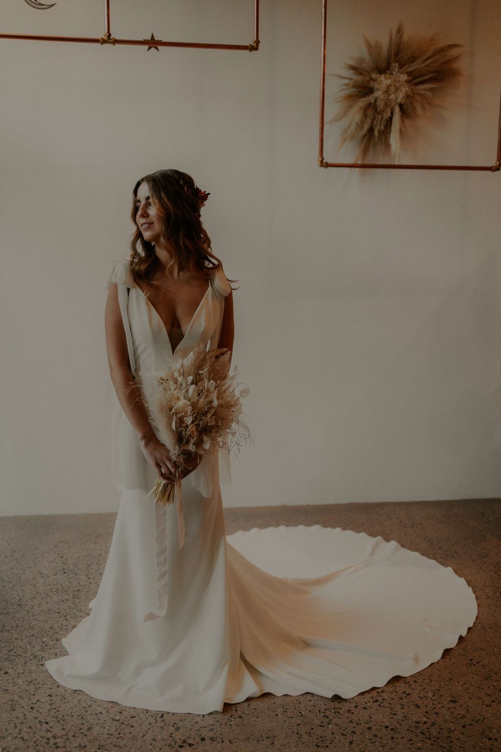 Elegant bride in flowing white dress with train holding dried flower bouquet against minimal wall decor.