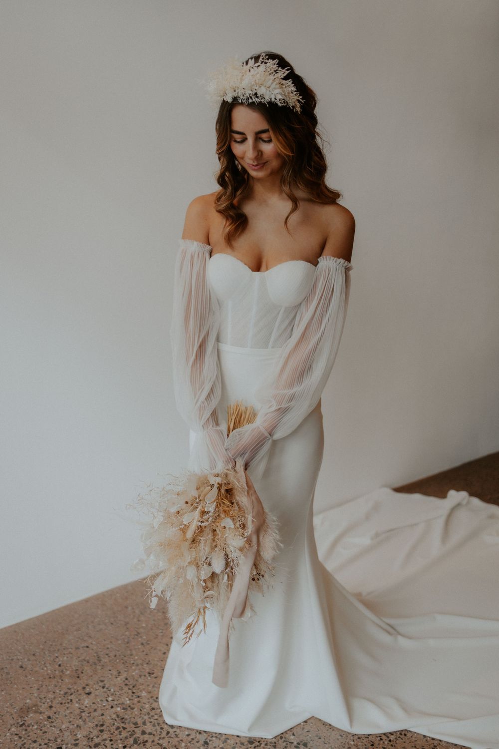 Elegant mermaid wedding dress with sheer sleeves and dried floral bouquet in studio setting.