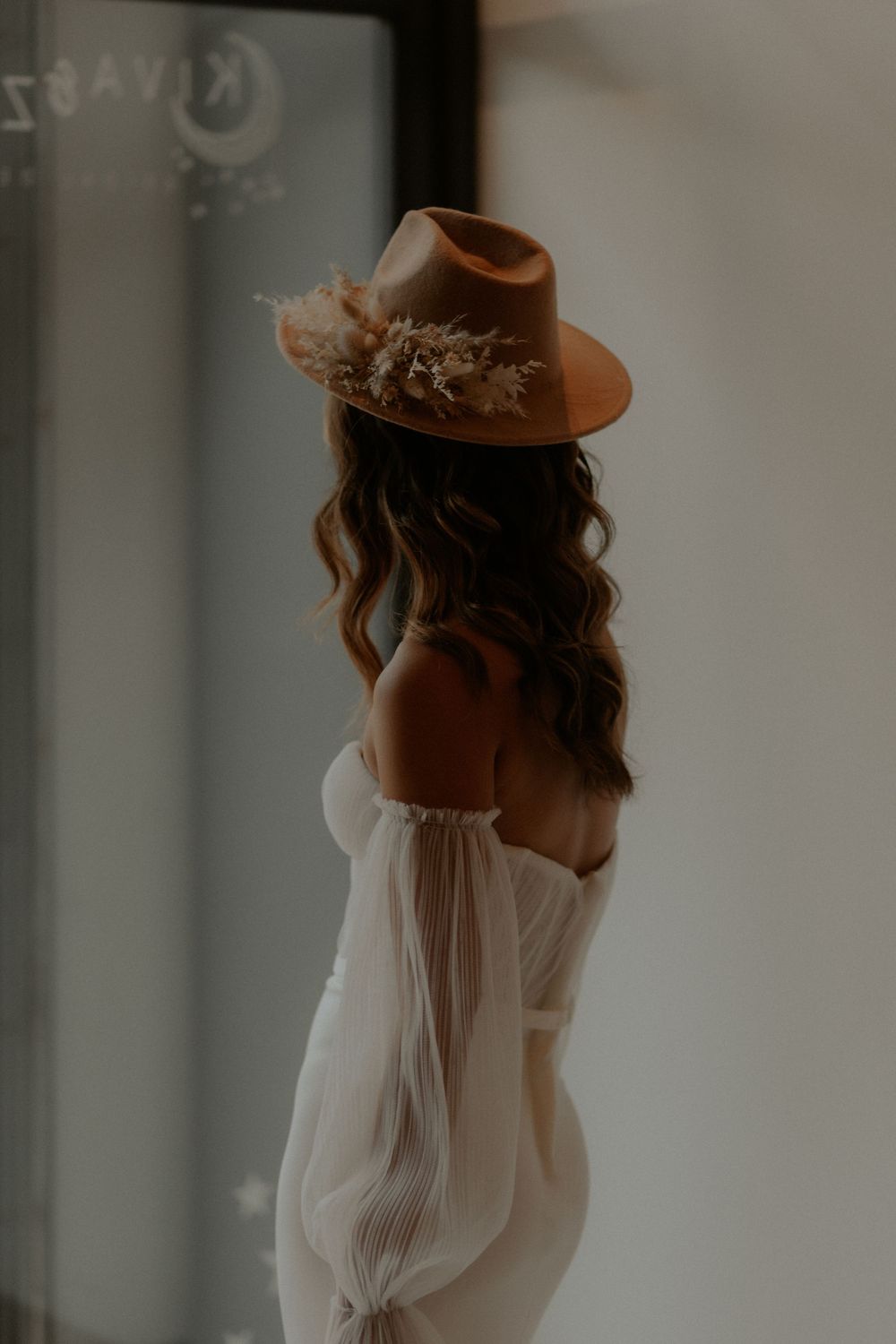 Romantic wedding portrait featuring an off-shoulder white dress and brown hat adorned with dried flowers.