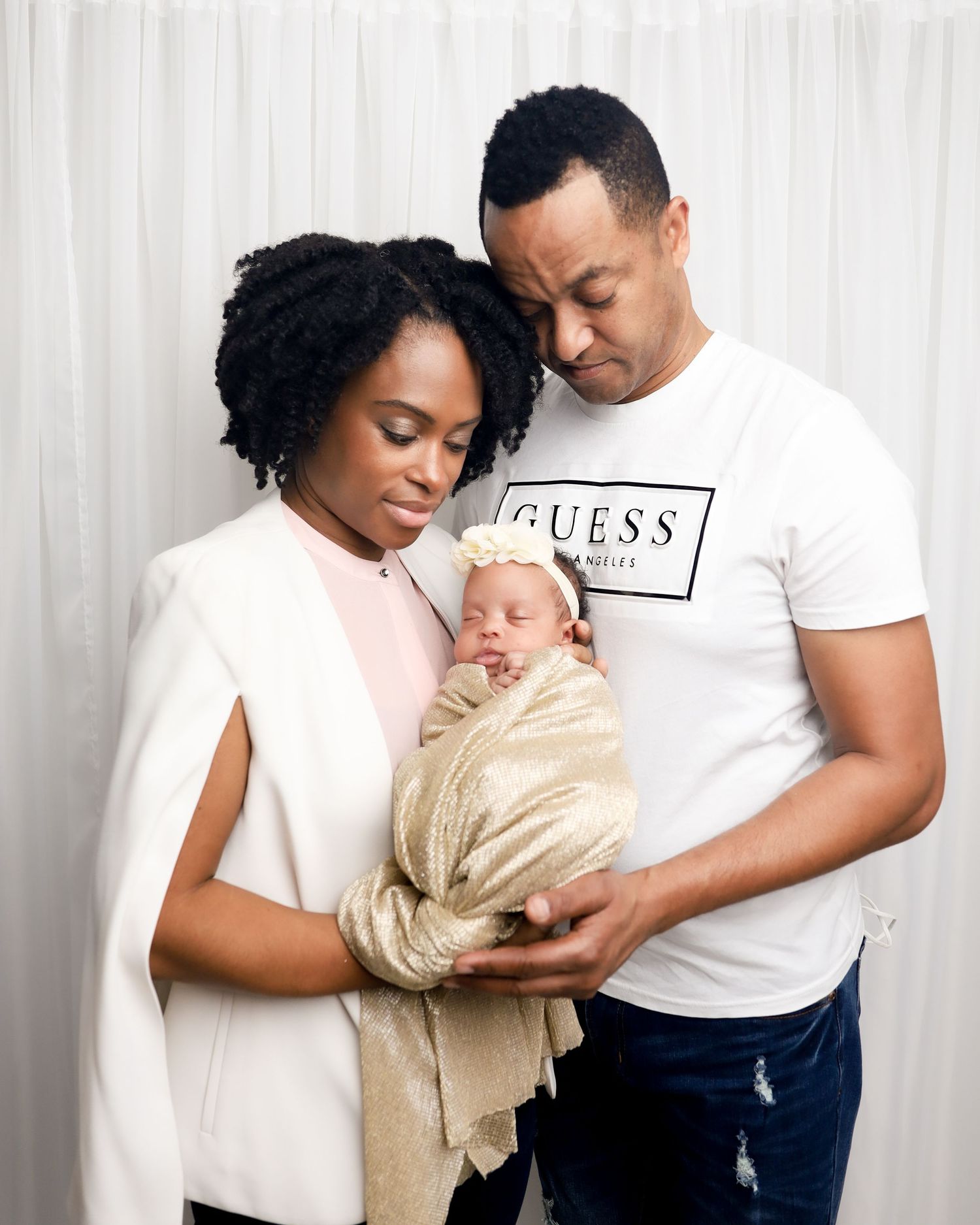 Two adults holding a sleeping newborn baby wrapped in beige fabric against a white backdrop.
