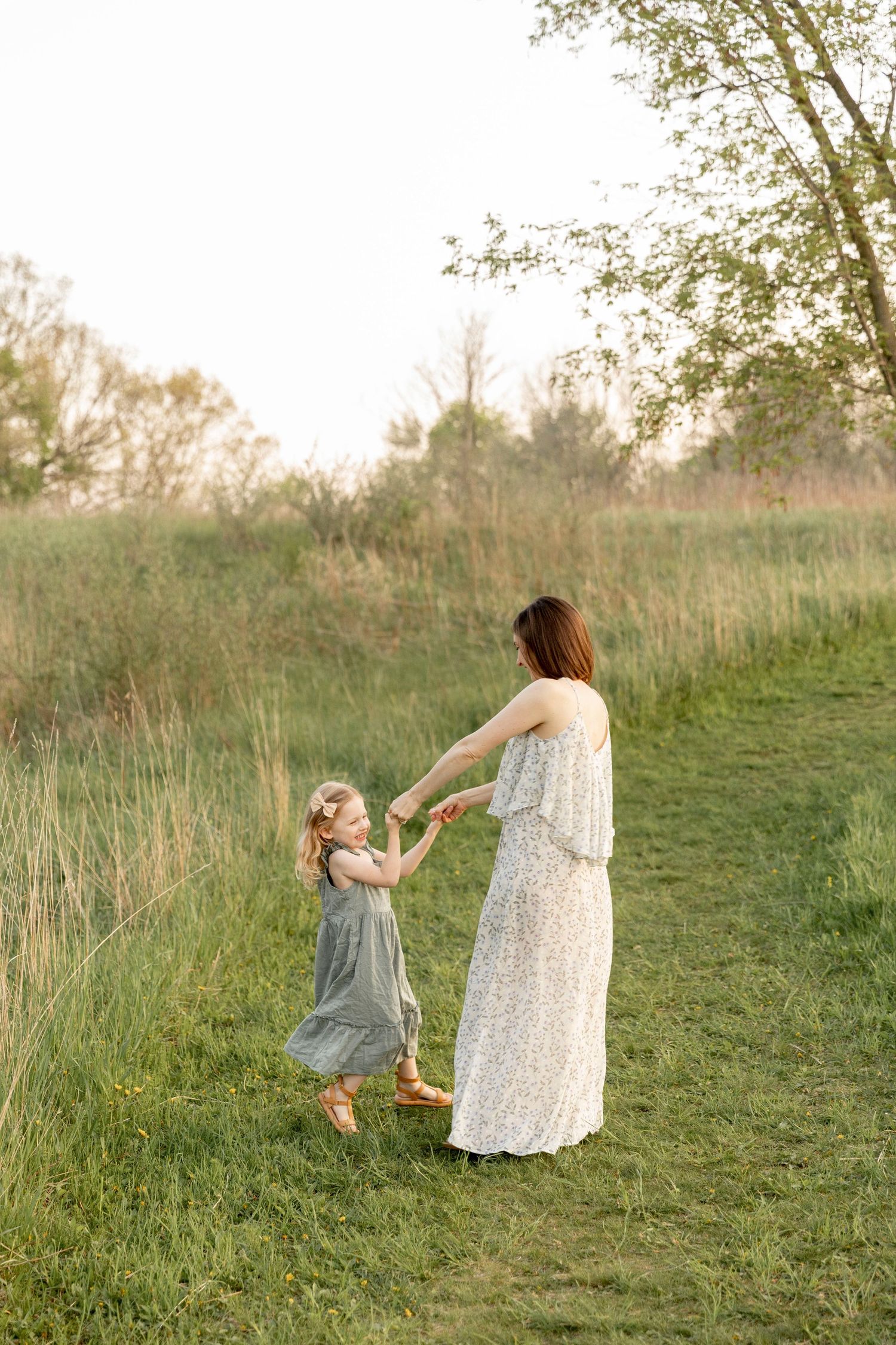 a mother and daughter in flowy dresses dance together in grasses during golden hour at bear creek nature preserve in Rochester Michigan