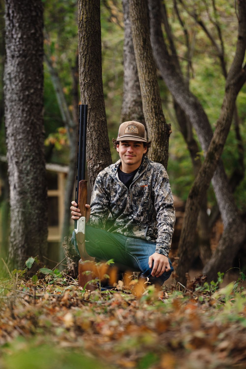 A hunter in camouflage gear kneels in autumn woods with a rifle.