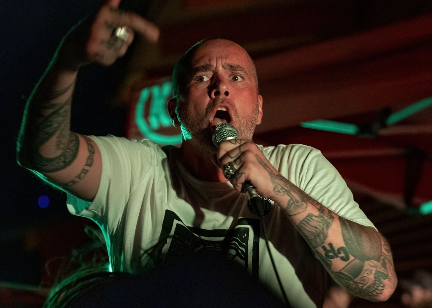 Energetic performer with tattoos singing passionately into microphone under green stage lighting.
