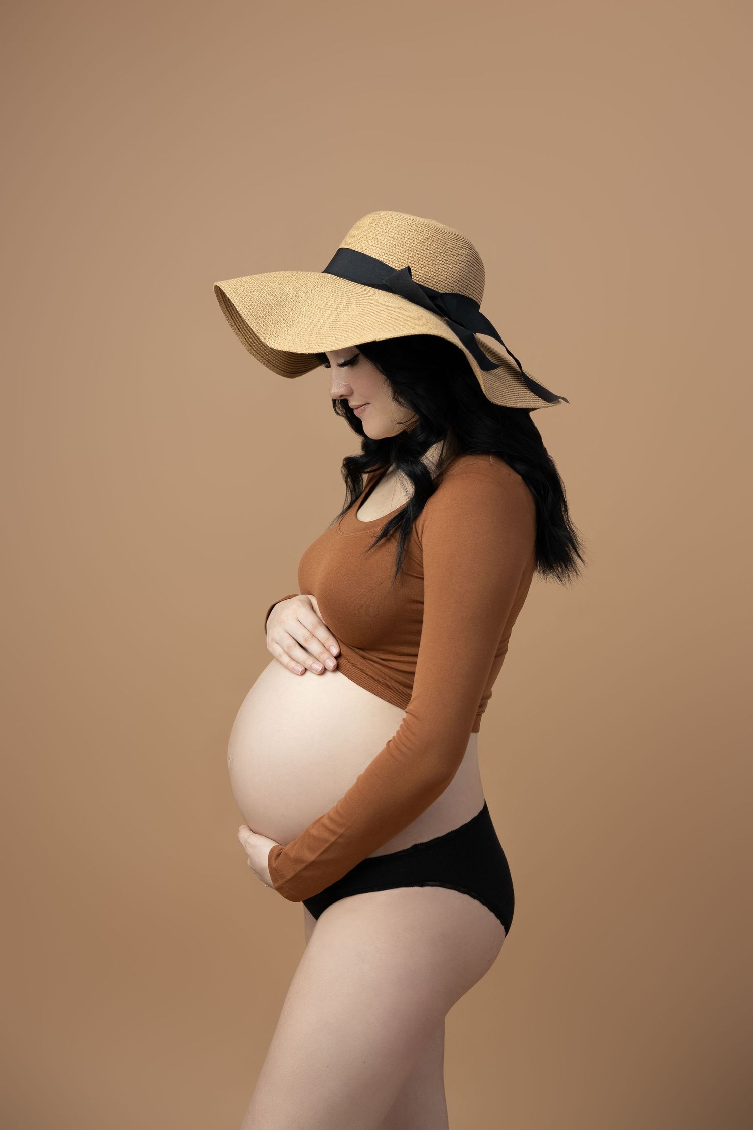 Maternity portrait with sun hat and brown outfit against beige backdrop.