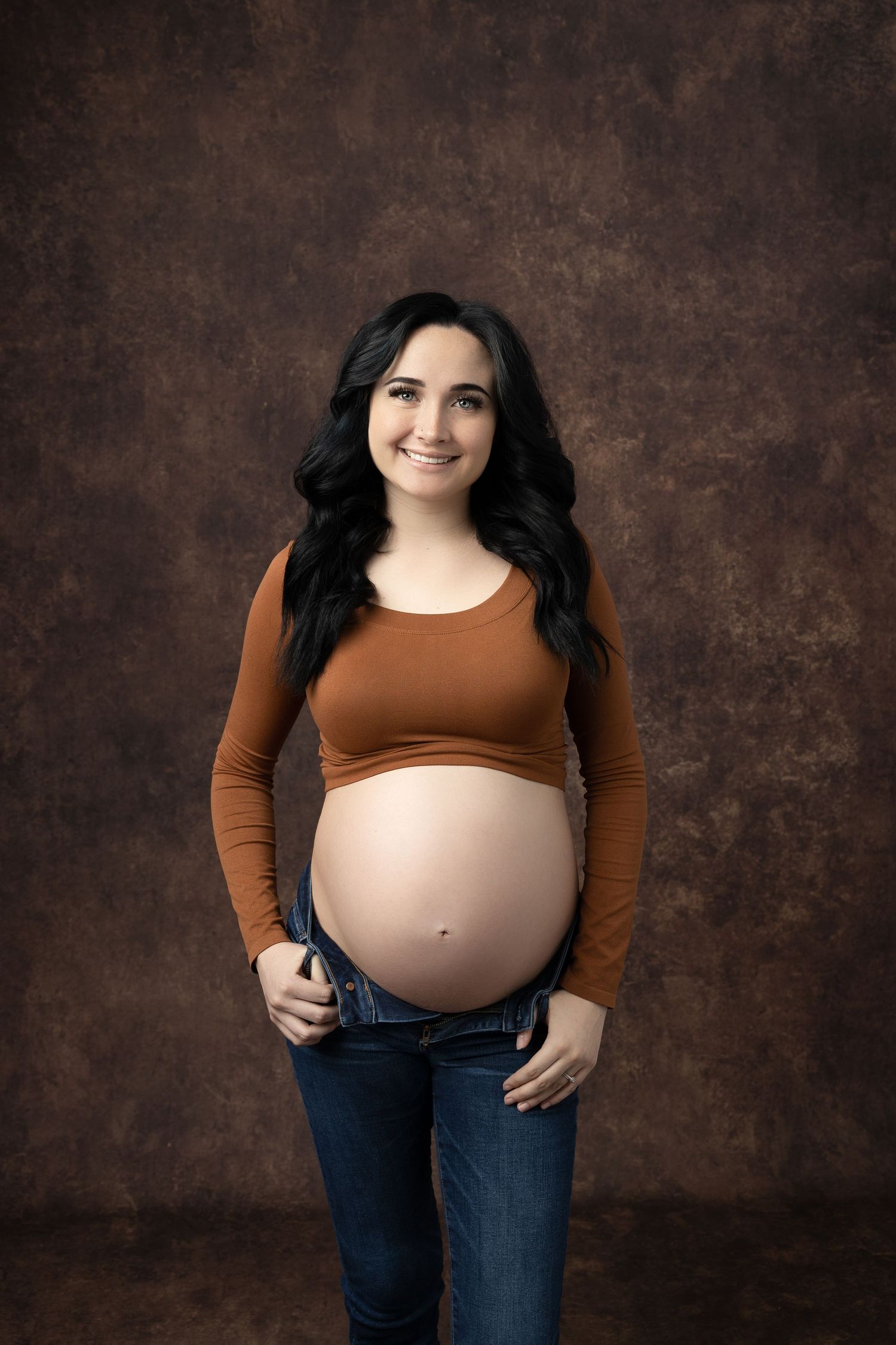 Maternity studio portrait with brown top and jeans against textured backdrop.