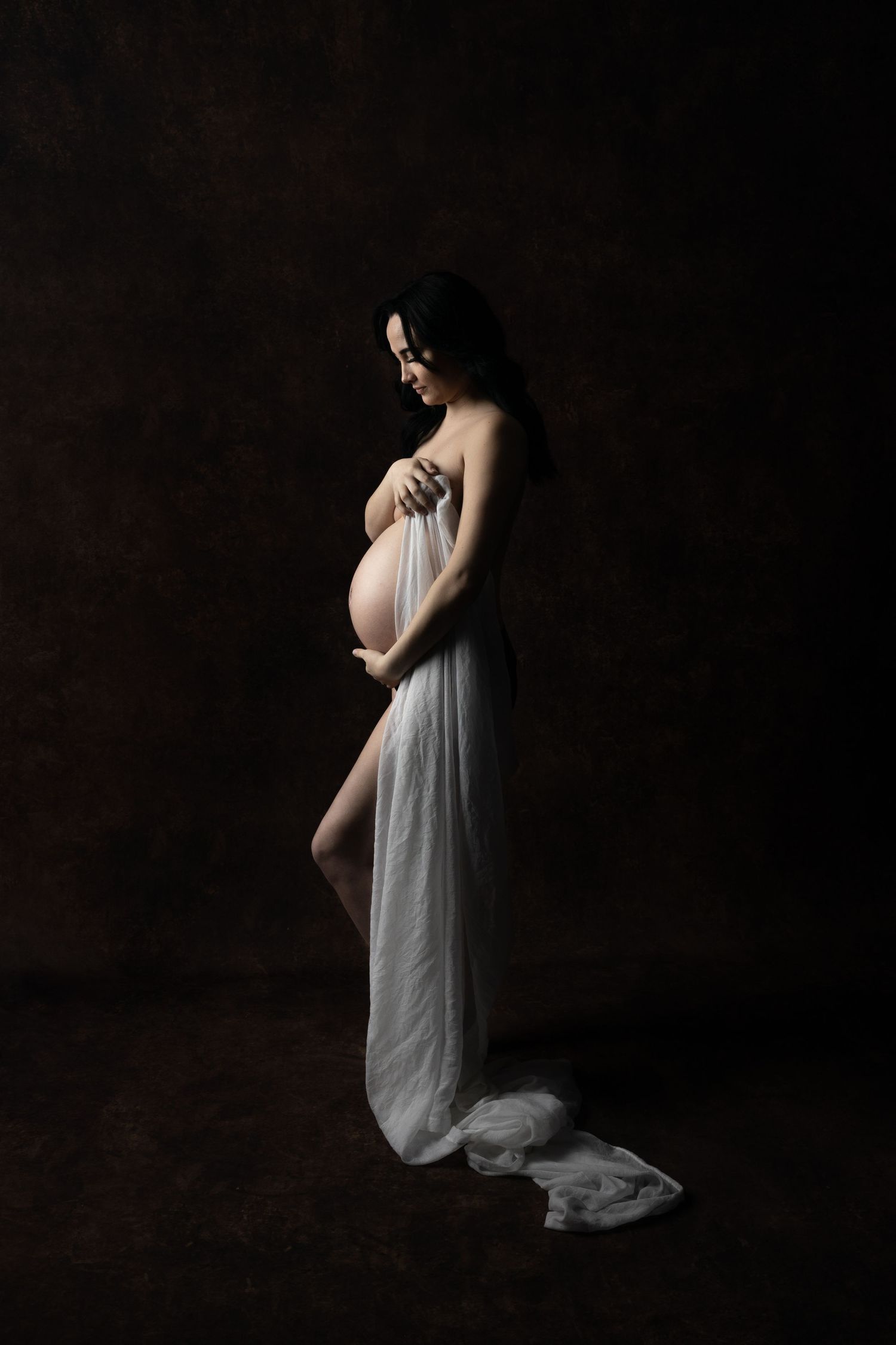 Dramatic silhouette maternity photos with flowing grey fabric in dark studio setting.