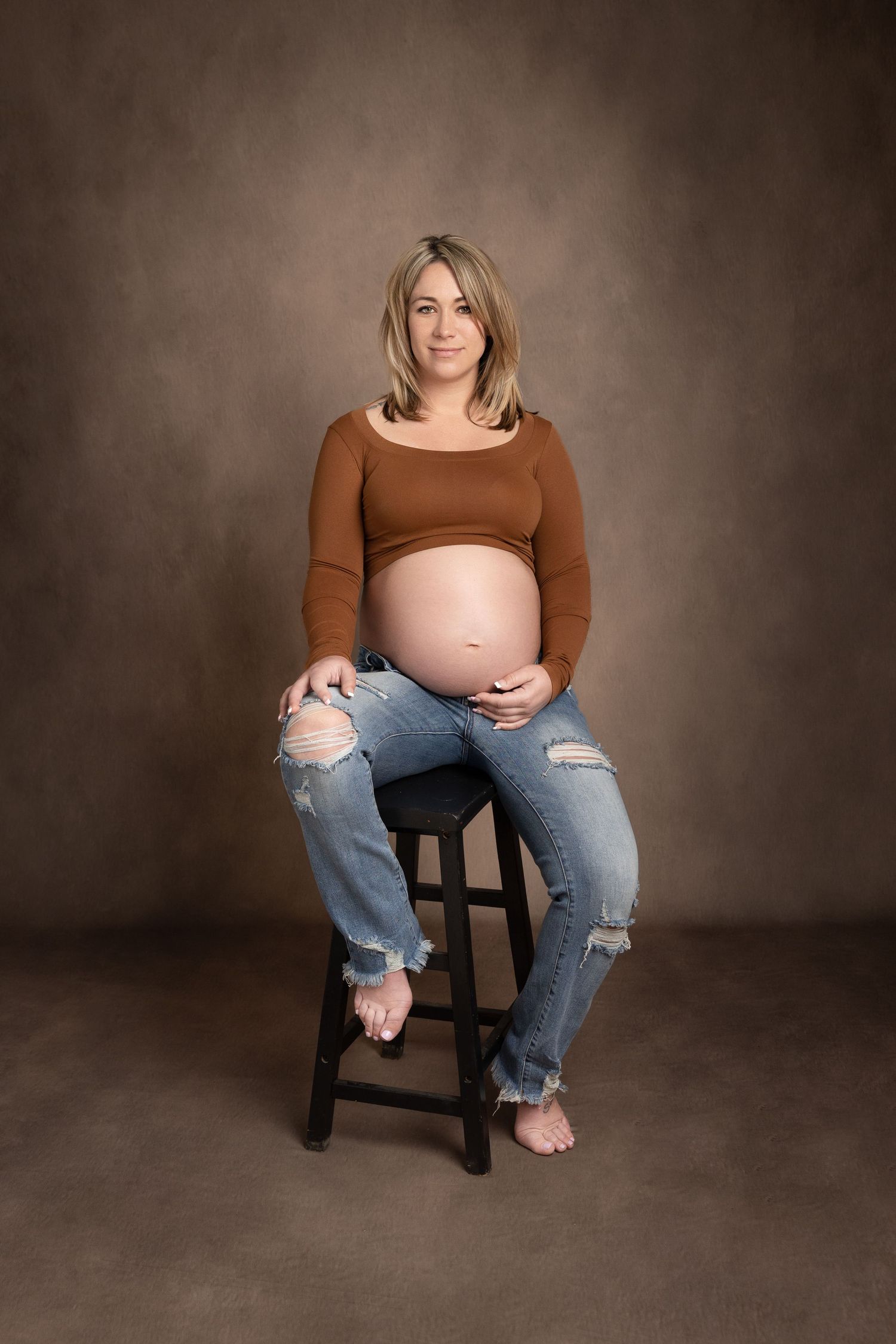 Casual maternity session featuring distressed jeans and brown top against moody backdrop.