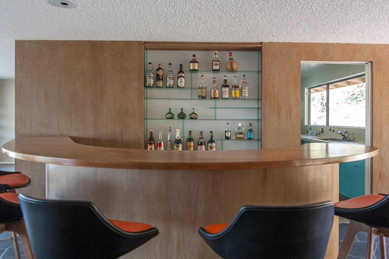 Modern wooden bar counter with built-in shelving displaying liquor bottles and black leather barstools.