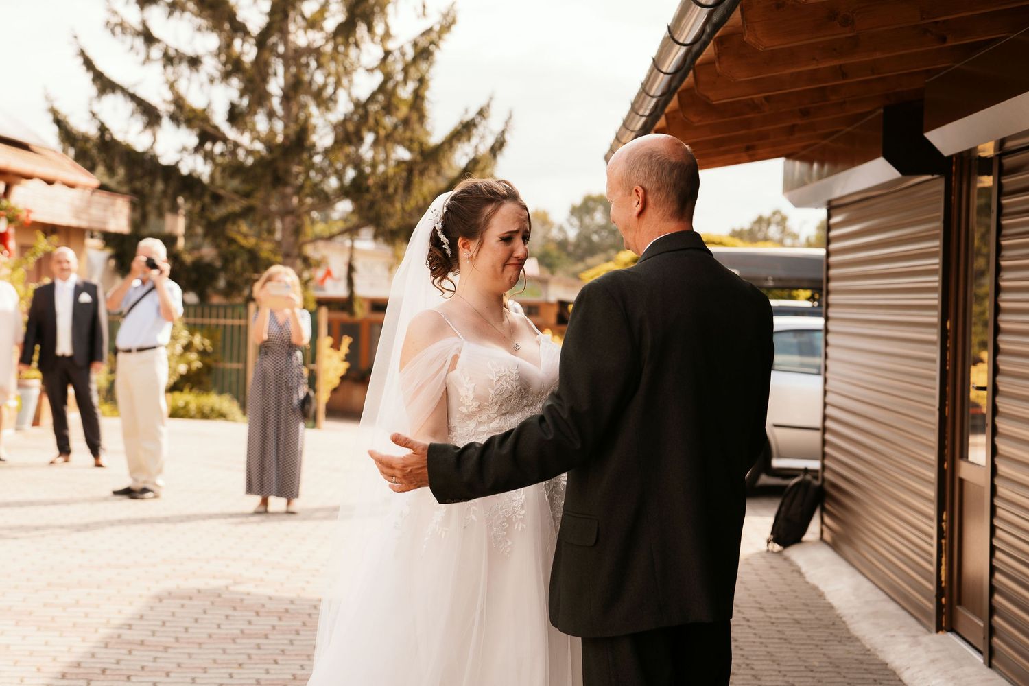 A bride and groom share their first look outside a building with guests in the background.