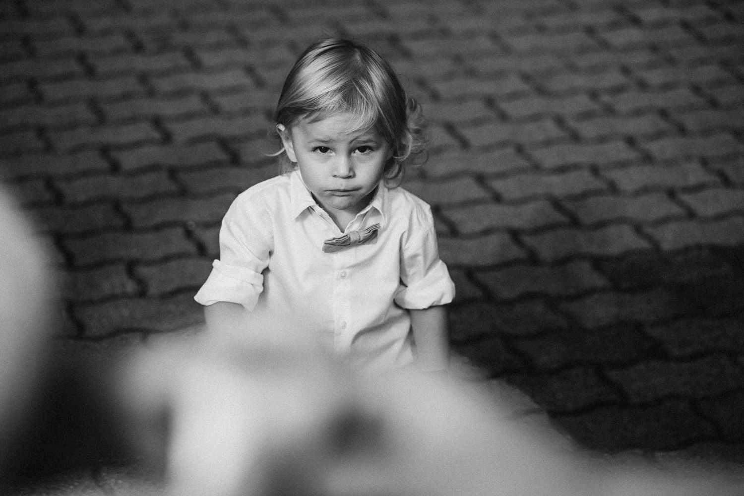 A black and white portrait of a young child sitting on cobblestones with a contemplative expression.