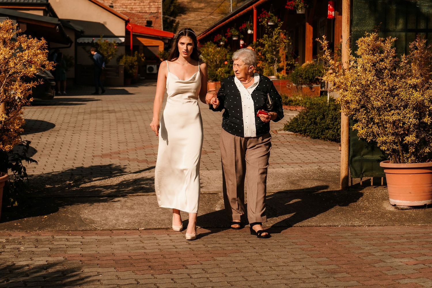 Two people walking together along a brick pathway surrounded by autumn foliage and potted plants.