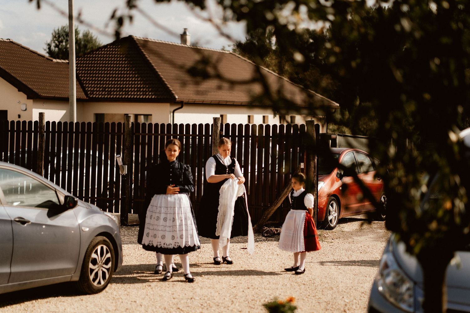 Three people in traditional dress standing near parked cars on a residential street.