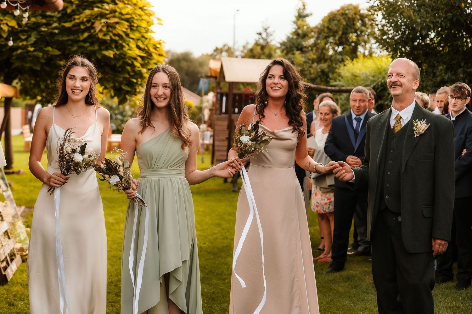 Wedding party members in elegant dresses standing together on a sunlit lawn.