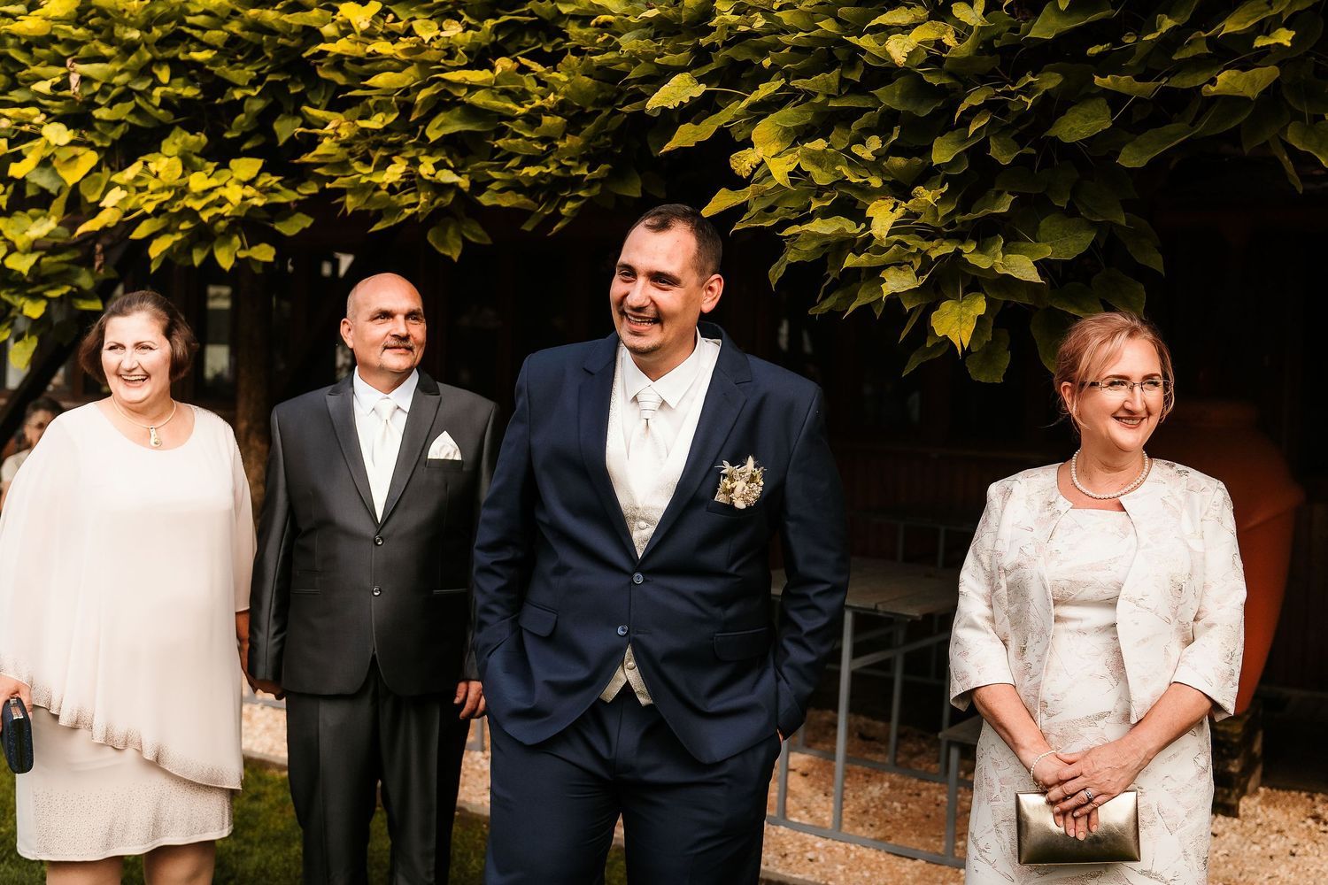Wedding party members smiling and standing together in formal attire against green foliage.