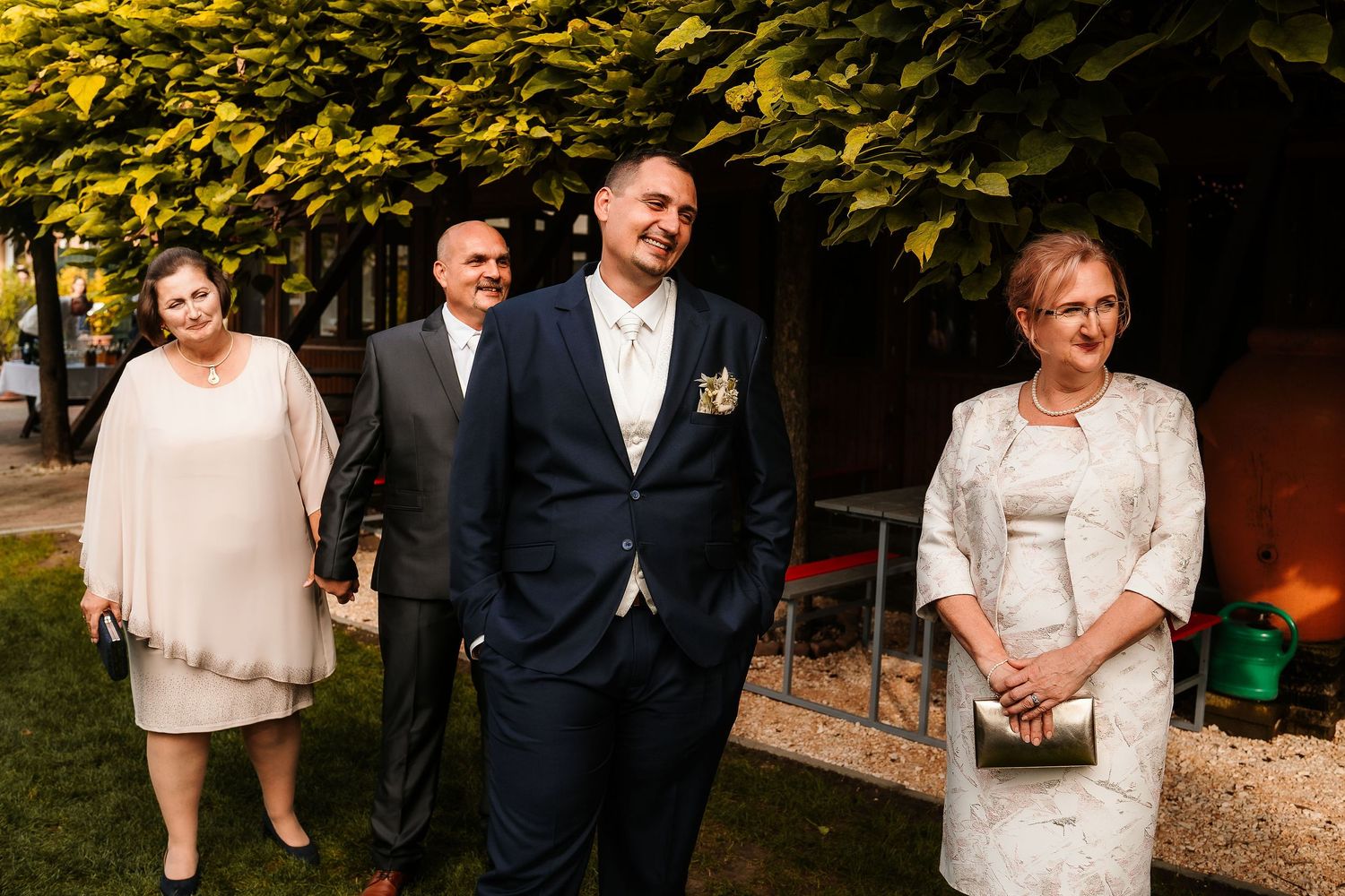 Wedding party members in formal attire standing together against green foliage.