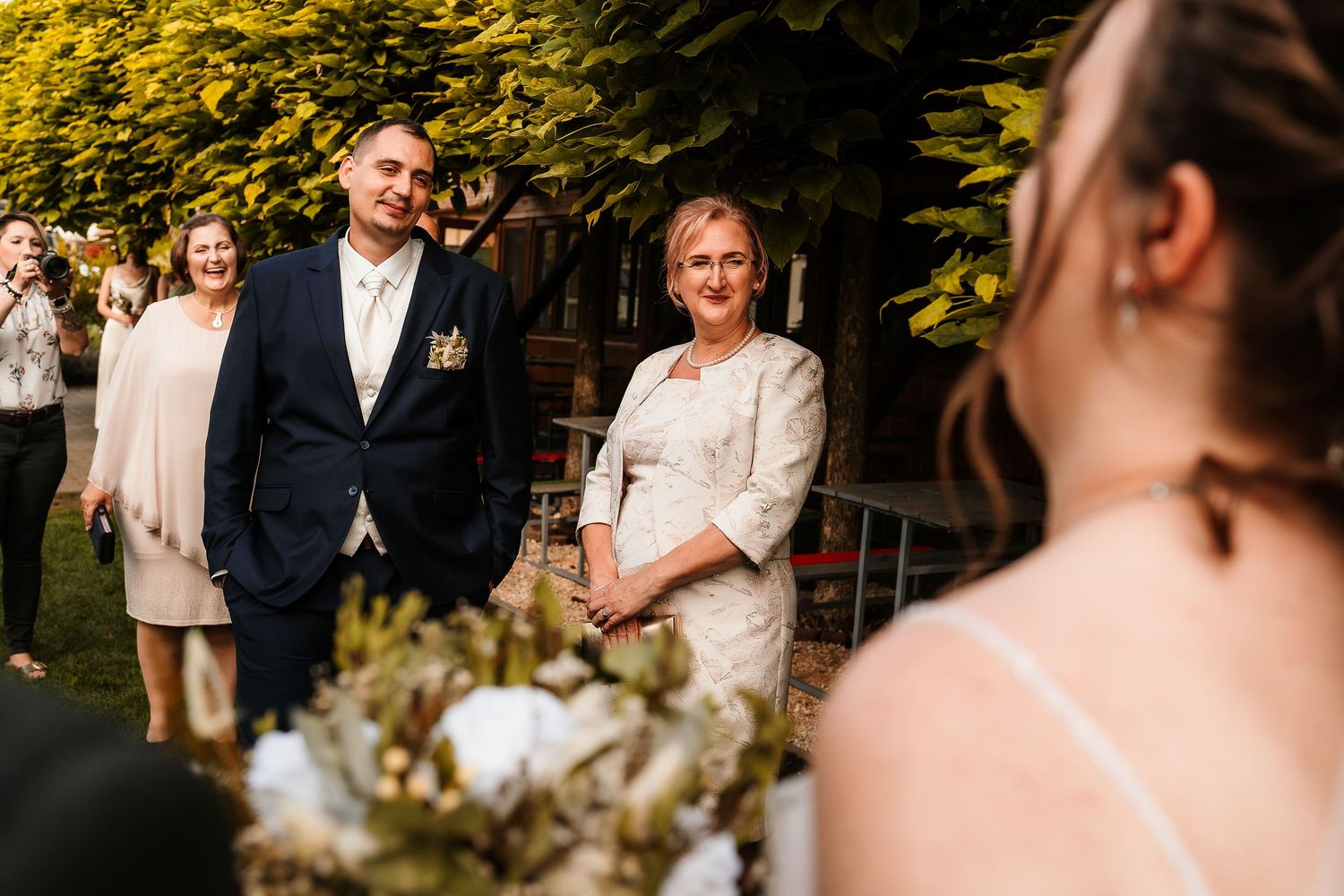 An emotional scene at an outdoor wedding ceremony with guests gathered under golden sunlit trees.