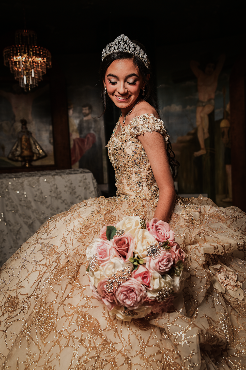 An ornate quinceañera dress with beaded details and a pink rose bouquet in a dramatic indoor setting.