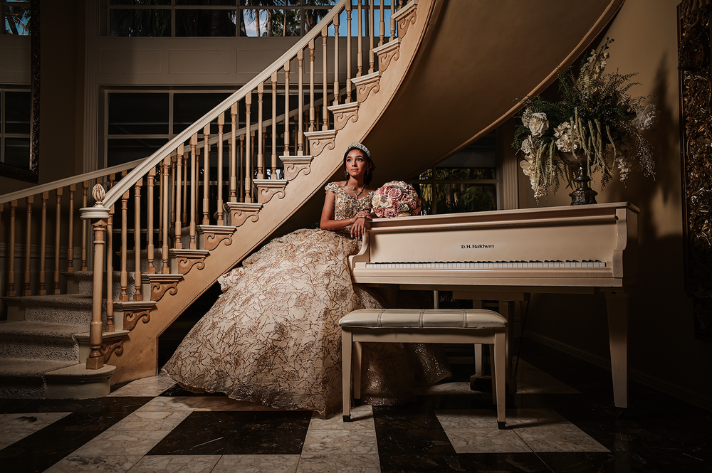 An elegant portrait by a grand piano and wooden staircase featuring a champagne-colored ballgown.